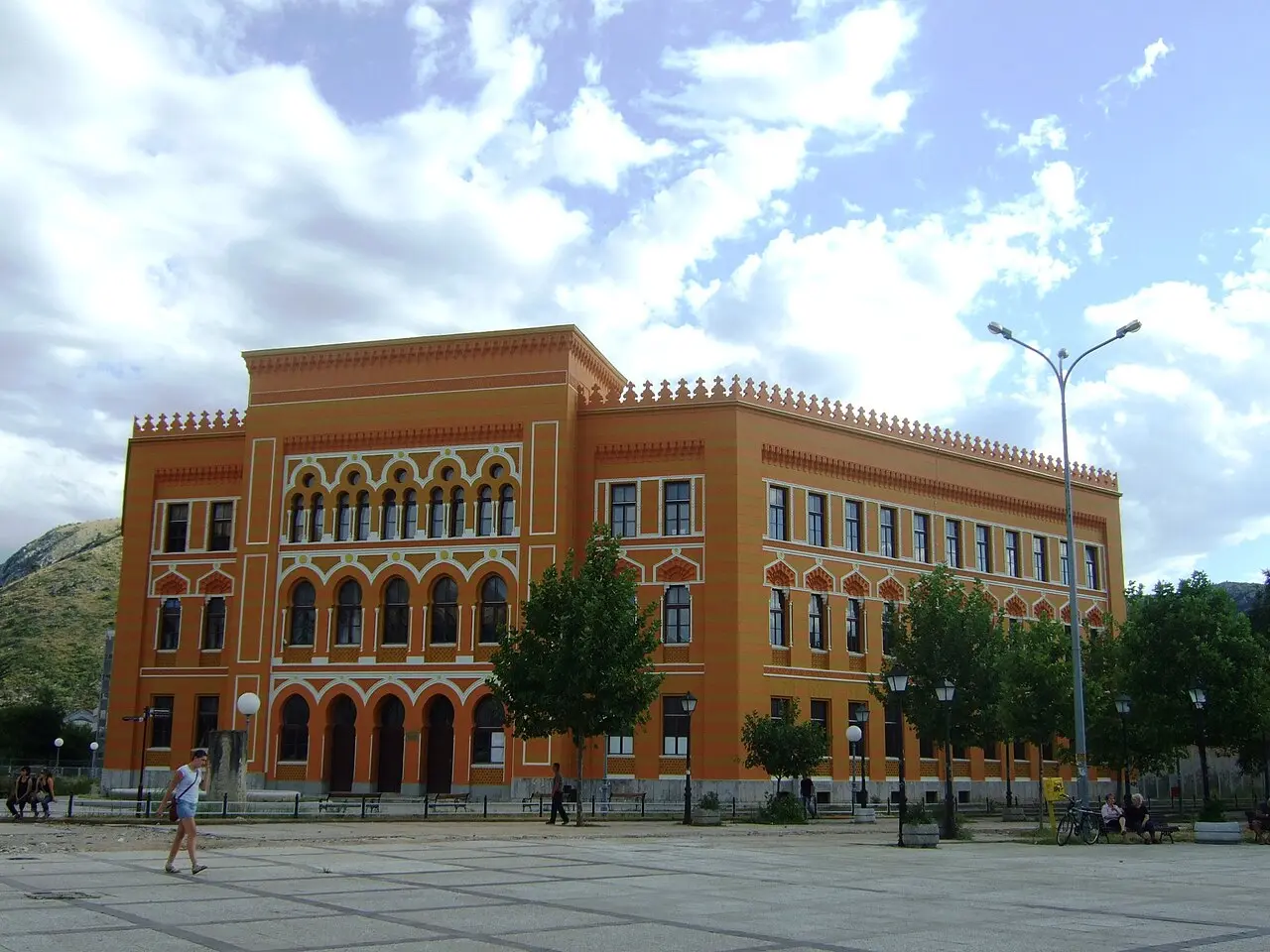 The restored orange Old Gymnasium building on the wide open Spanish Square on the modern west bank of Mostar