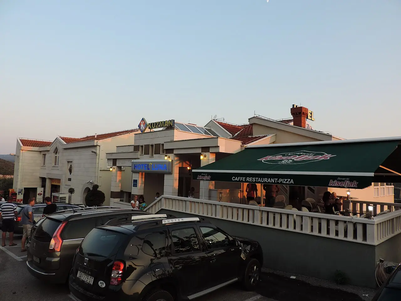 A small hotel and a cafe restaurant with a terrace and parked cars along a street in Neum at dusk
