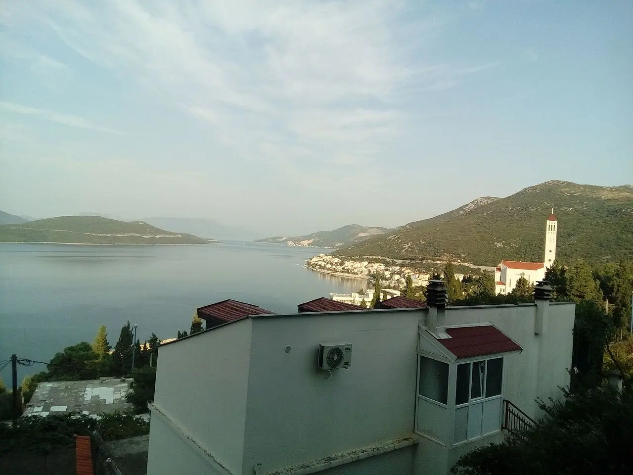 View over Neum bay from an apartment terrace at dawn, with a church bell tower and the town along the far shore