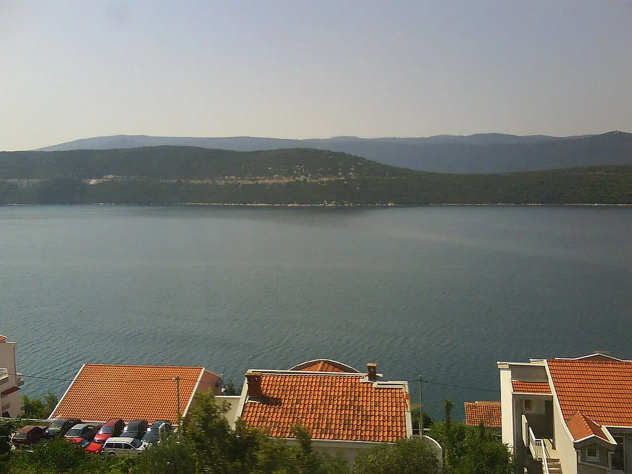 Red-roofed apartment houses on the Neum hillside above the calm bay with cars parked below