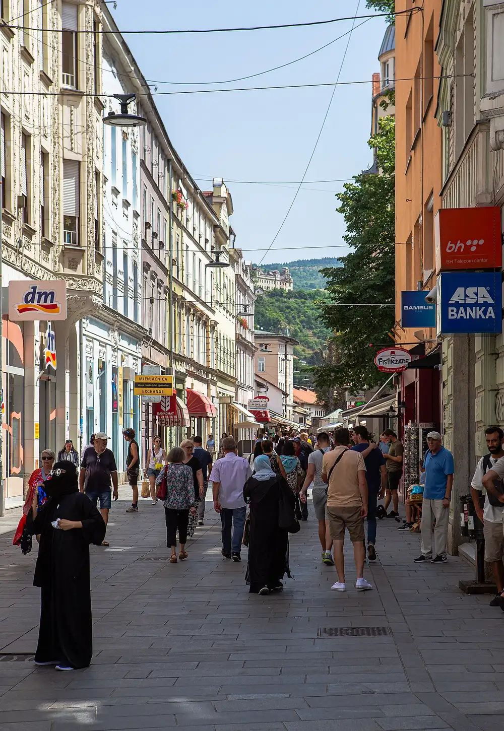 The pedestrian Ferhadija street in Sarajevo busy with people in summer, Austro-Hungarian buildings and shops, the green Trebević hill closing the view