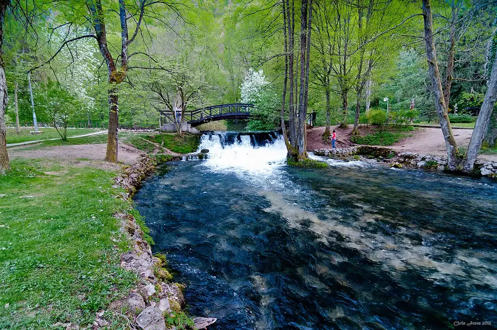 The Vrelo Bosne springs park at Ilidža, a clear river running over a low cascade under a wooden footbridge among green trees