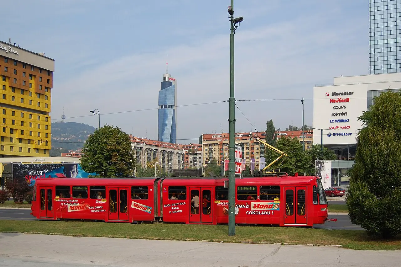 A red Sarajevo tram signed for line 3 Baščaršija, with the Avaz Twist Tower and Holiday hotel behind at Marijin Dvor