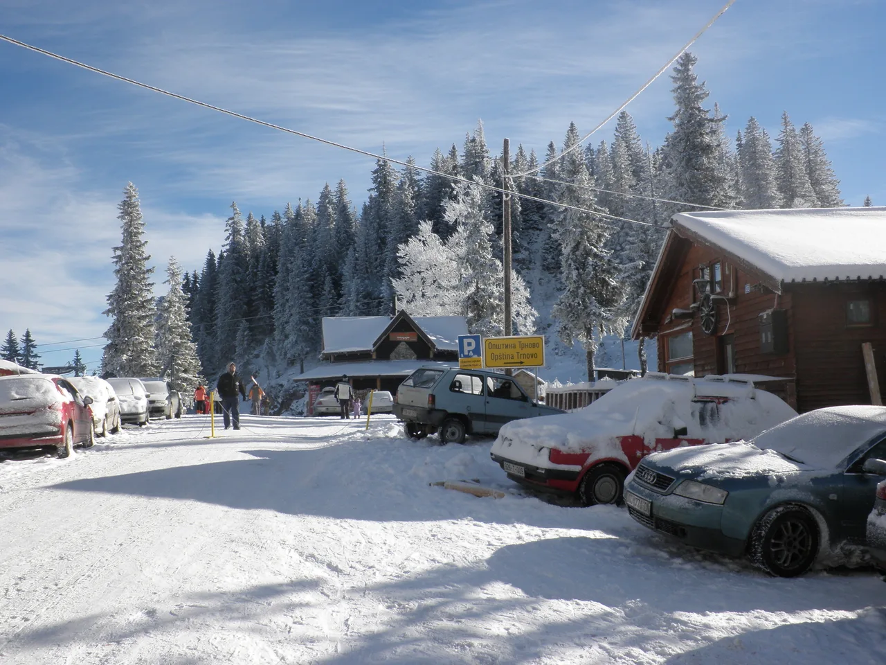 Cars parked along the snowy resort road on Jahorina beside a parking sign and timber buildings