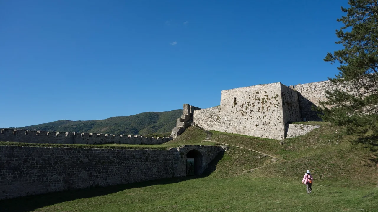 The grassy inner courtyard and stone ramparts of Jajce fortress on a clear day, wooded hills rising behind the walls