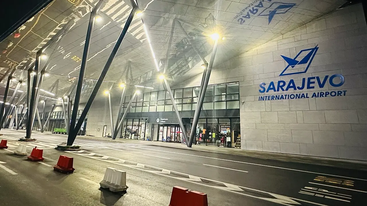 The illuminated terminal facade of Sarajevo International Airport at night, with the SARAJEVO INTERNATIONAL AIRPORT sign lit up