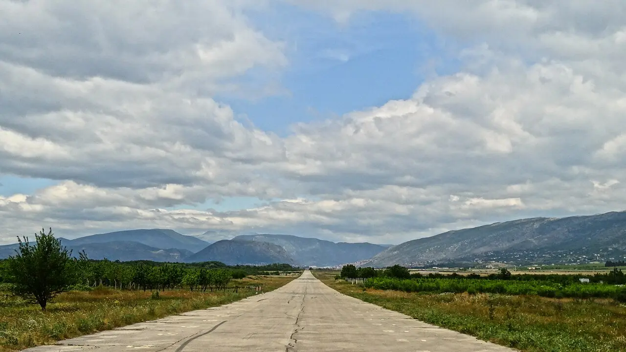 The quiet approach road to Mostar International Airport running straight toward the Herzegovina mountains under a cloudy sky