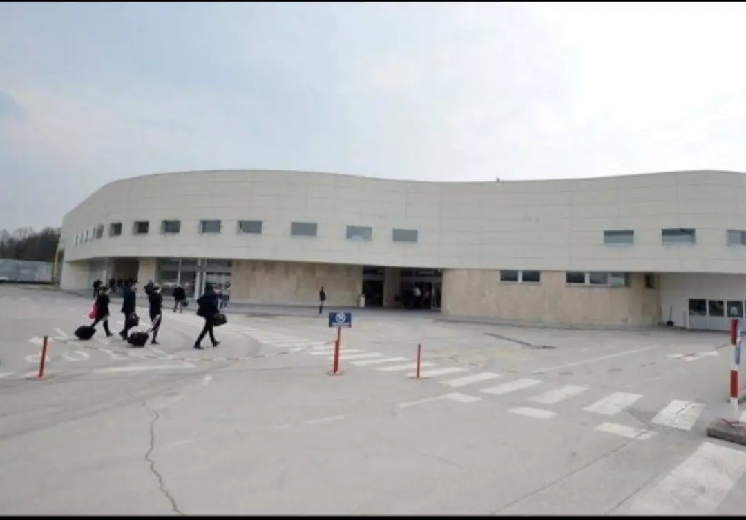 Passengers wheeling luggage across the forecourt of the low white terminal at Tuzla International Airport, the budget gateway in north-east Bosnia