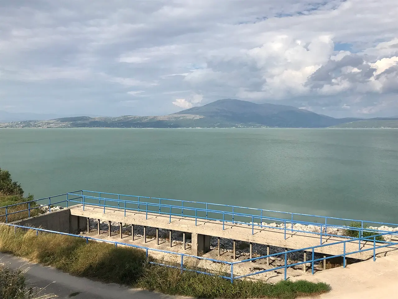 The wide pale-green expanse of Busko lake under a cloudy sky, a low mountain ridge on the far shore and a concrete overlook with blue railings in front