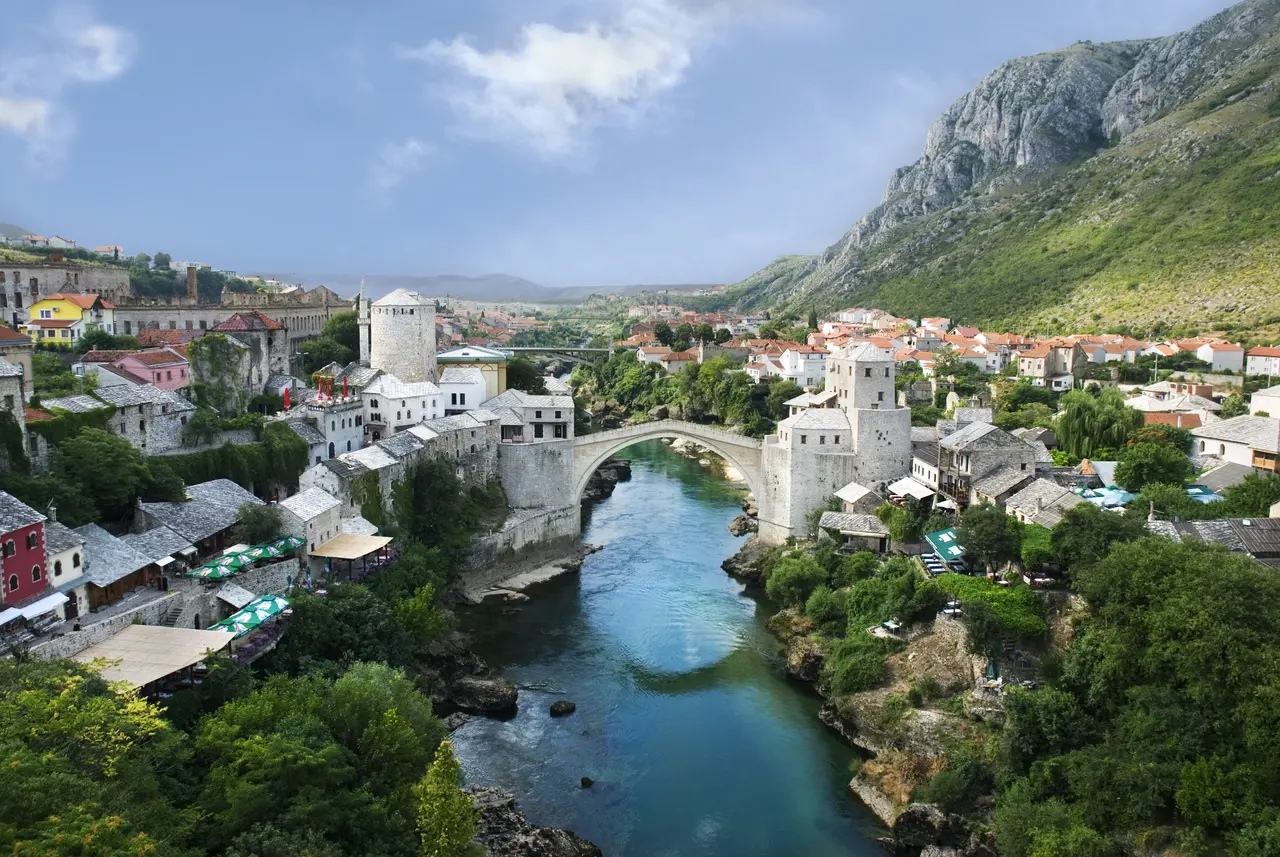 Wide panorama of Mostar old town: the stone arch of Stari Most, the bazaar roofs, a mosque minaret and the green Neretva with bare hills beyond